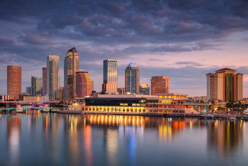 Tampa skyline is bathed in the soft, golden light of sunrise. Skyscrapers reflect off the calm waters, highlighting modern architecture with varied heights and designs. The sky transitions from warm hues near the horizon to deeper blues above, with scattered clouds adding texture. The waterfront is illuminated, casting colorful reflections that enhance the serene atmosphere. The scene captures a tranquil yet dynamic urban landscape, embodying the city's vibrant morning energy.
